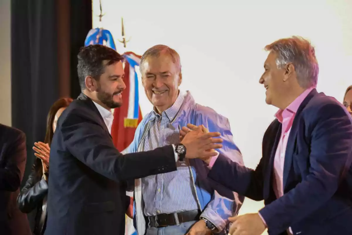 Three men smile and greet each other enthusiastically at a formal event, while other people applaud in the background. Three men smile and greet each other enthusiastically at a formal event, while other people applaud in the background.