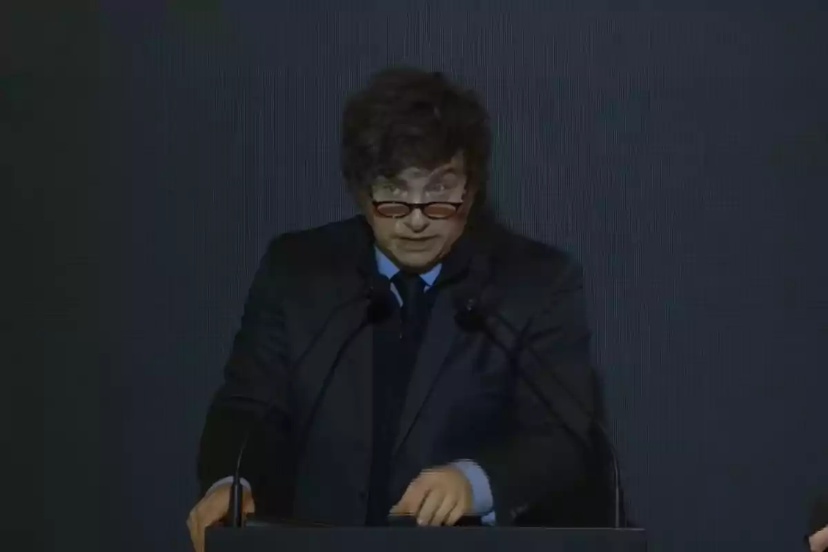 Dark-haired man with glasses speaking at a podium with a dark gray background