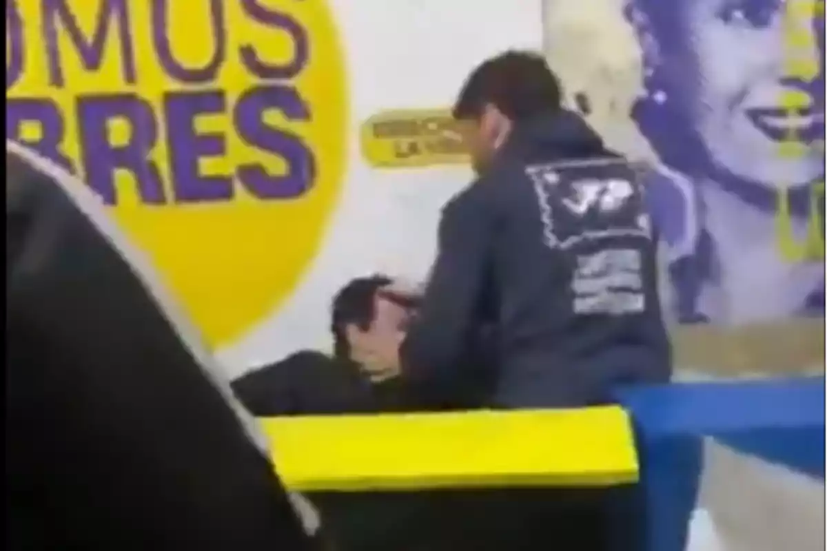 Un hombre ayuda a otra persona que está sentada frente a un mural con letras grandes y una imagen de una mujer sonriente al fondo.