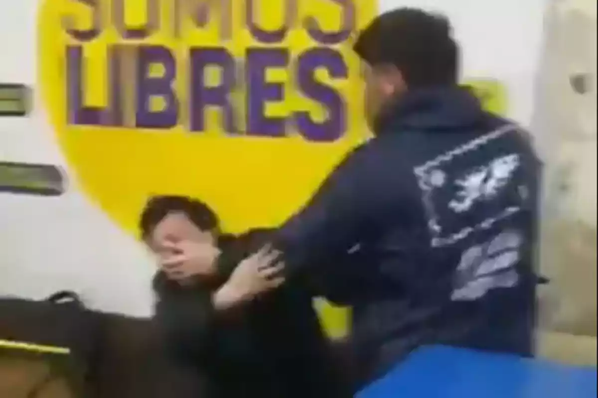 Un hombre con chaqueta oscura cubre su rostro mientras otro hombre lo sujeta frente a una pared amarilla con letras grandes que dicen somos libres Un hombre con chaqueta oscura cubre su rostro mientras otro hombre lo sujeta frente a una pared amarilla con letras grandes que dicen somos libres