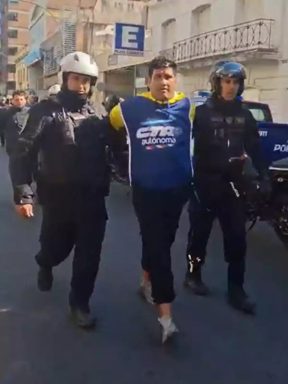 Un hombre con chaleco azul es escoltado por dos policías con uniforme negro y casco en una calle urbana. Un hombre con chaleco azul es escoltado por dos policías con uniforme negro y casco en una calle urbana.
