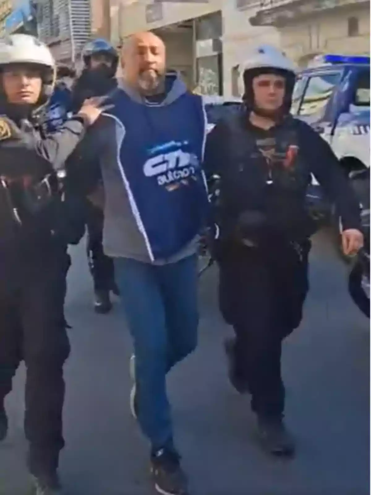 Un hombre con chaleco azul es escoltado por dos policías con casco en una calle urbana Un hombre con chaleco azul es escoltado por dos policías con casco en una calle urbana