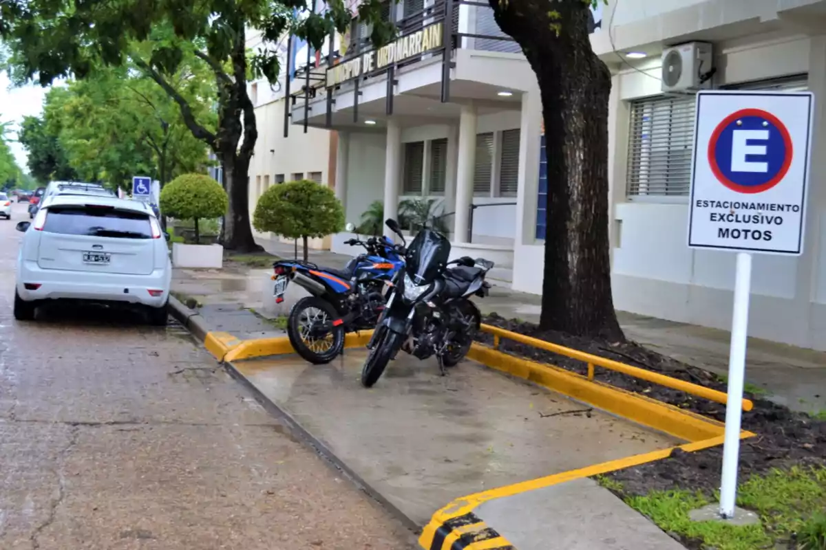Two motorcycles parked in a space reserved for motorcycles next to the sidewalk in front of a building with trees and a sign indicating the parking area. Two motorcycles parked in a space reserved for motorcycles next to the sidewalk in front of a building with trees and a sign indicating the parking area.