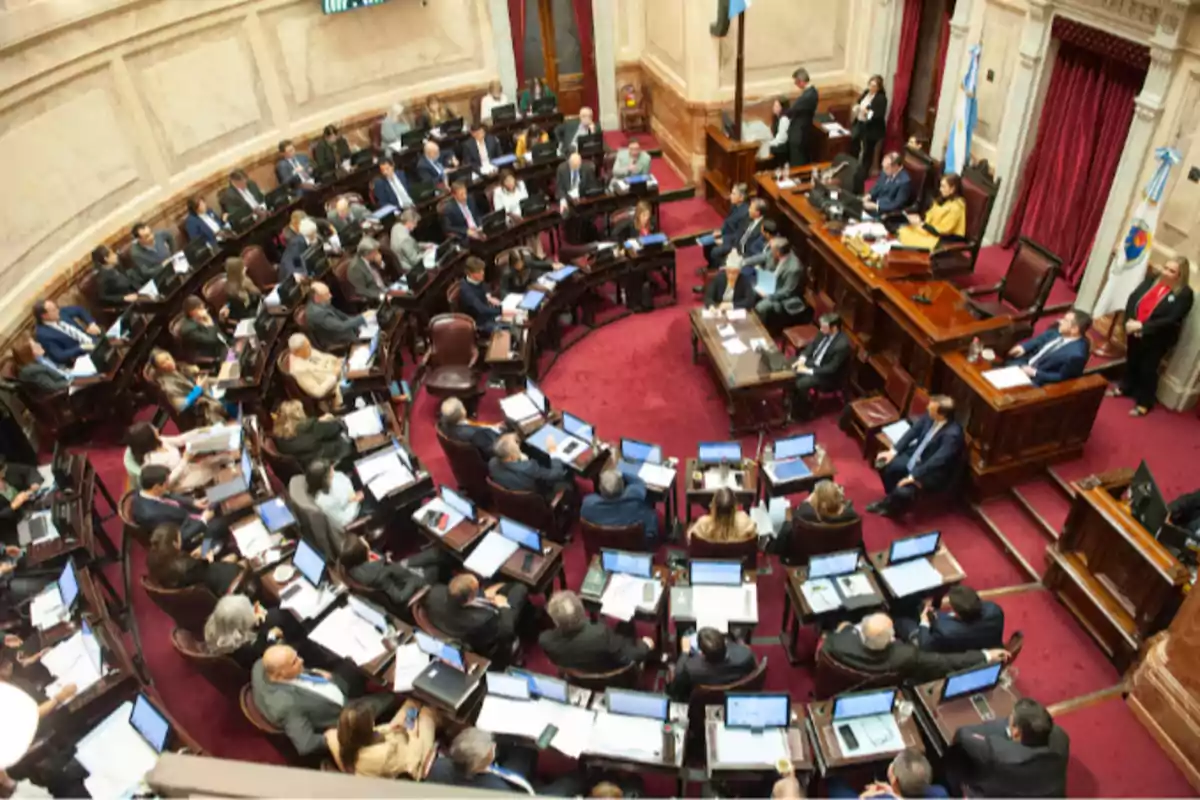 Top view of a parliamentary session with numerous legislators seated at their desks using laptops in a chamber with red carpeting