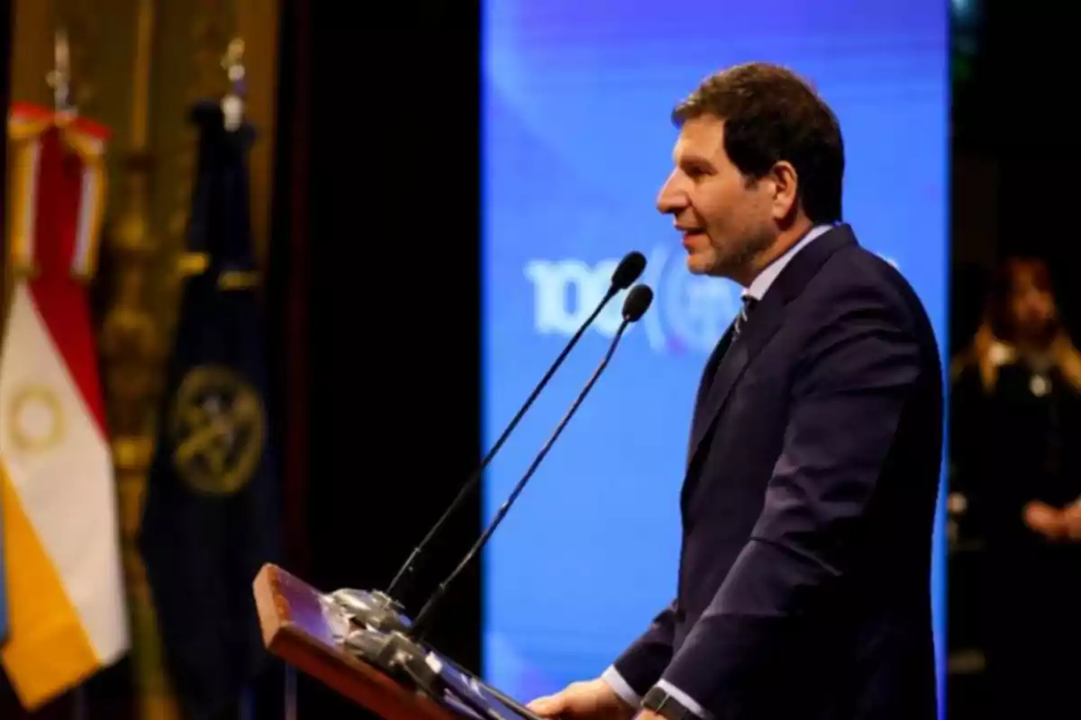 Man in a suit speaking at a podium with microphones in front of a blue screen and flags in the background Man in a suit speaking at a podium with microphones in front of a blue screen and flags in the background