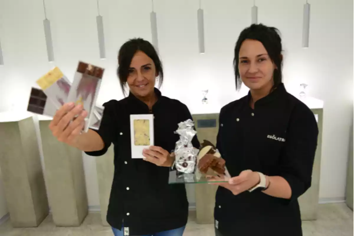 Dos mujeres con uniforme negro sostienen tabletas de chocolate y postres en un entorno moderno y minimalista
