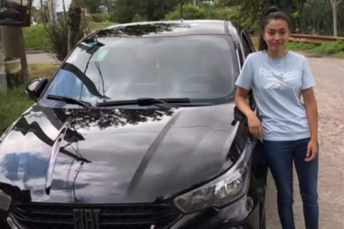 Una mujer joven con camiseta azul claro y jeans está de pie junto a un automóvil negro marca Fiat en un entorno al aire libre. Una mujer joven con camiseta azul claro y jeans está de pie junto a un automóvil negro marca Fiat en un entorno al aire libre.