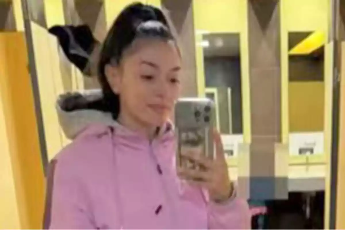 Joven con sudadera rosa tomándose una selfie frente a un espejo en un baño público con paredes amarillas. Joven con sudadera rosa tomándose una selfie frente a un espejo en un baño público con paredes amarillas.