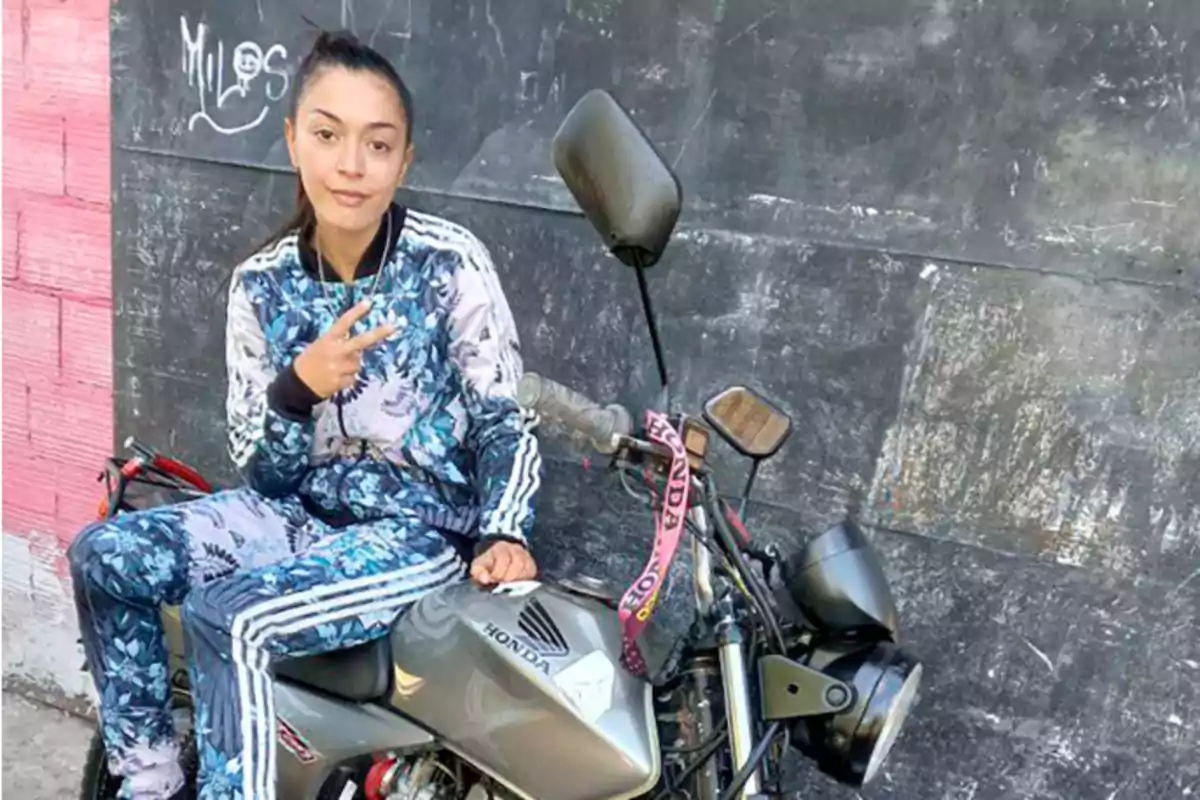 Joven con conjunto deportivo azul sentado en una motocicleta Honda haciendo la señal de paz frente a una pared oscura