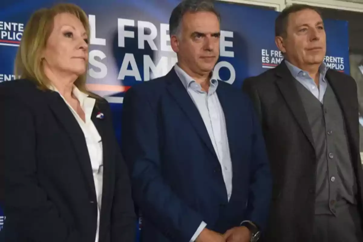 Tres personas vestidas de manera formal posan frente a un cartel azul que dice "El Frente es Amplio"