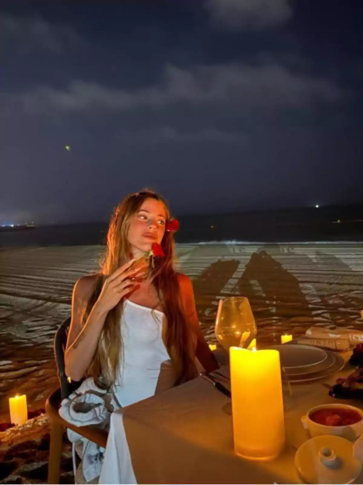 Mujer con vestido blanco sentada en una mesa decorada con velas en la playa durante la noche sosteniendo una flor roja