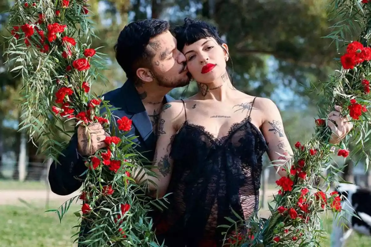 Pareja con tatuajes posando juntos en un columpio decorado con flores rojas en un entorno natural