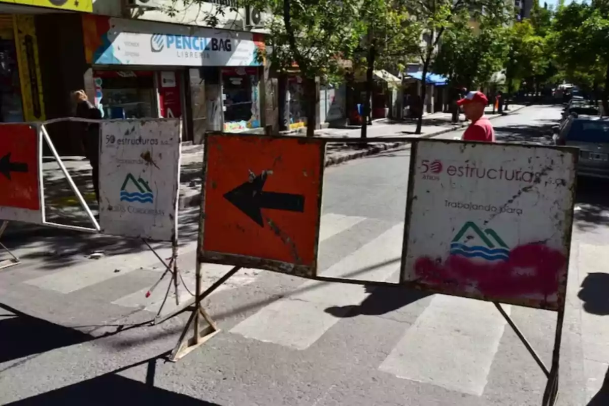 Barricadas metálicas con señales de desvío bloquean una calle en una zona urbana con árboles y comercios al fondo