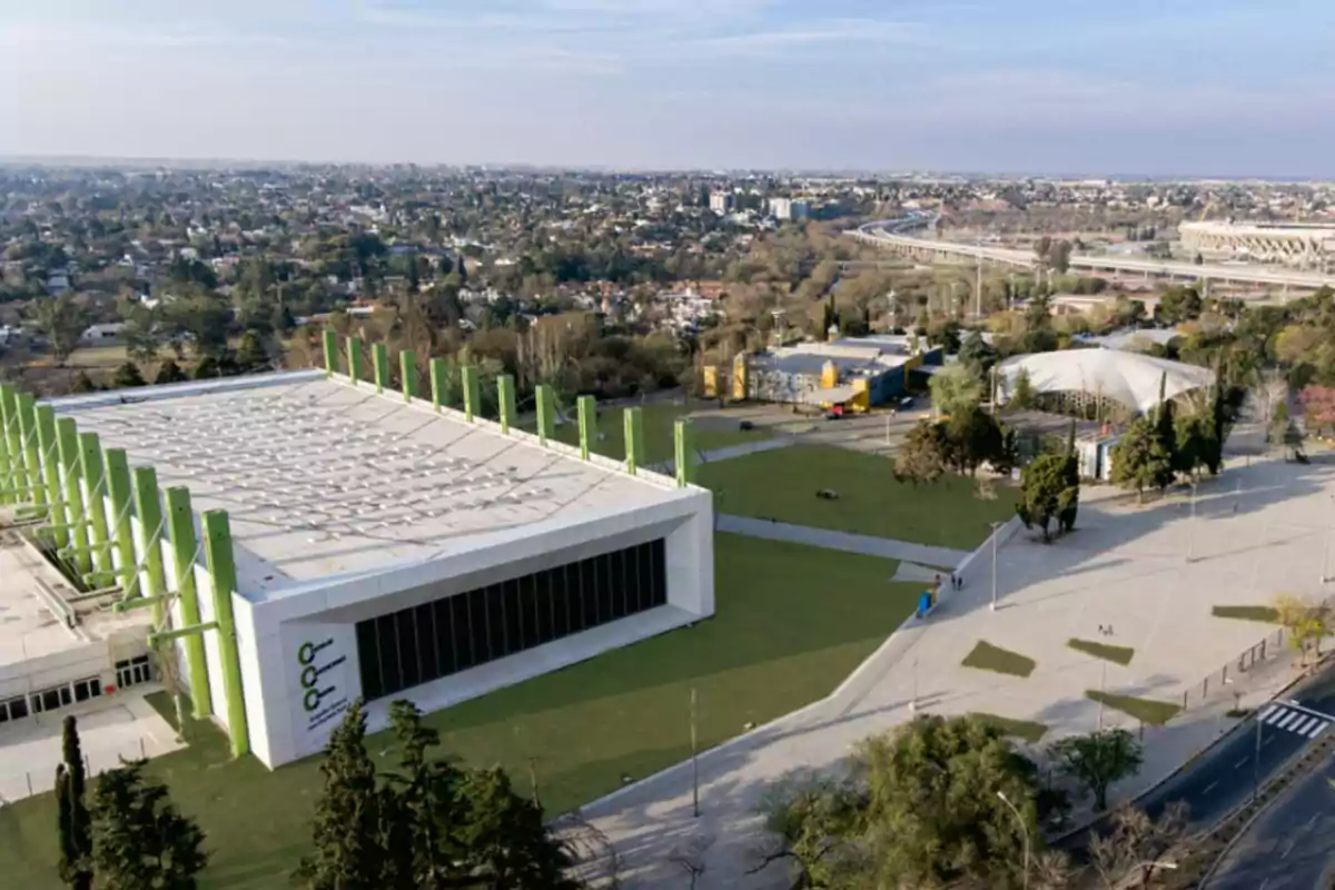 Aerial view of a large modern building with green columns, surrounded by green areas, pedestrian paths, and a city in the background.