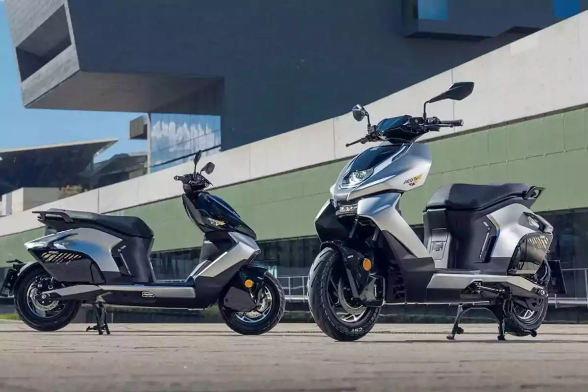 Dos motos tipo scooter eléctricas de color gris y negro están estacionadas al aire libre frente a un edificio moderno Dos motos tipo scooter eléctricas de color gris y negro están estacionadas al aire libre frente a un edificio moderno