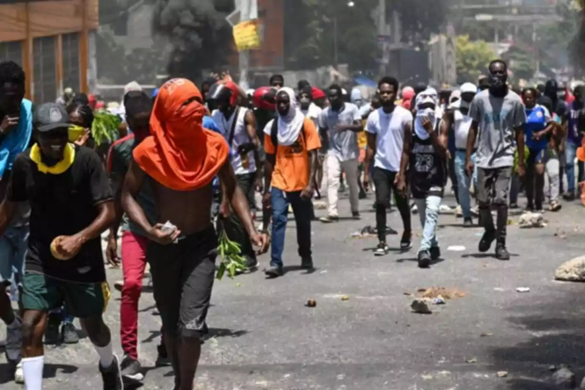 Un grupo de personas camina por una calle durante una protesta, algunas llevan el rostro cubierto y al fondo se observa humo negro.