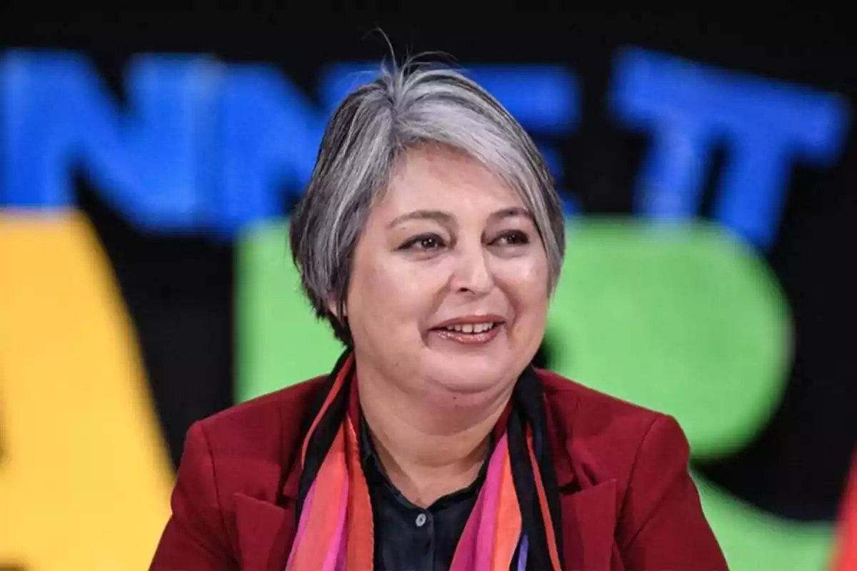 Mujer de cabello corto y canoso sonríe mientras viste saco rojo y bufanda colorida con fondo de letras grandes y coloridas desenfocadas Mujer de cabello corto y canoso sonríe mientras viste saco rojo y bufanda colorida con fondo de letras grandes y coloridas desenfocadas