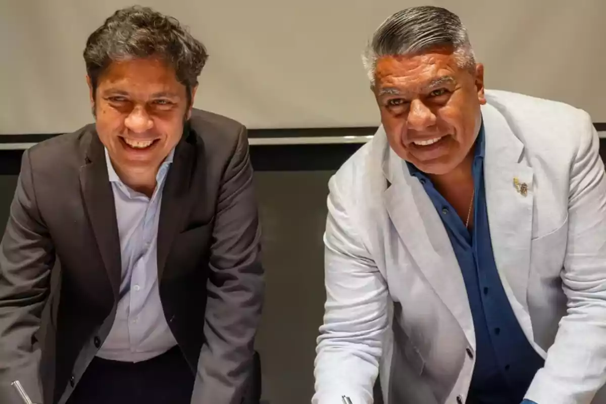 Dos hombres sonrientes vestidos con saco posan juntos inclinados hacia adelante en un ambiente interior iluminado. Dos hombres sonrientes vestidos con saco posan juntos inclinados hacia adelante en un ambiente interior iluminado.
