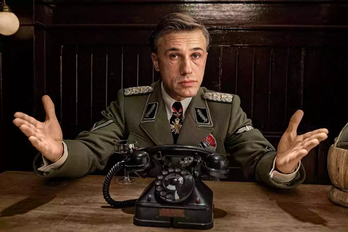 Hombre con uniforme militar sentado en una mesa de madera con un teléfono antiguo frente a él y los brazos extendidos Hombre con uniforme militar sentado en una mesa de madera con un teléfono antiguo frente a él y los brazos extendidos