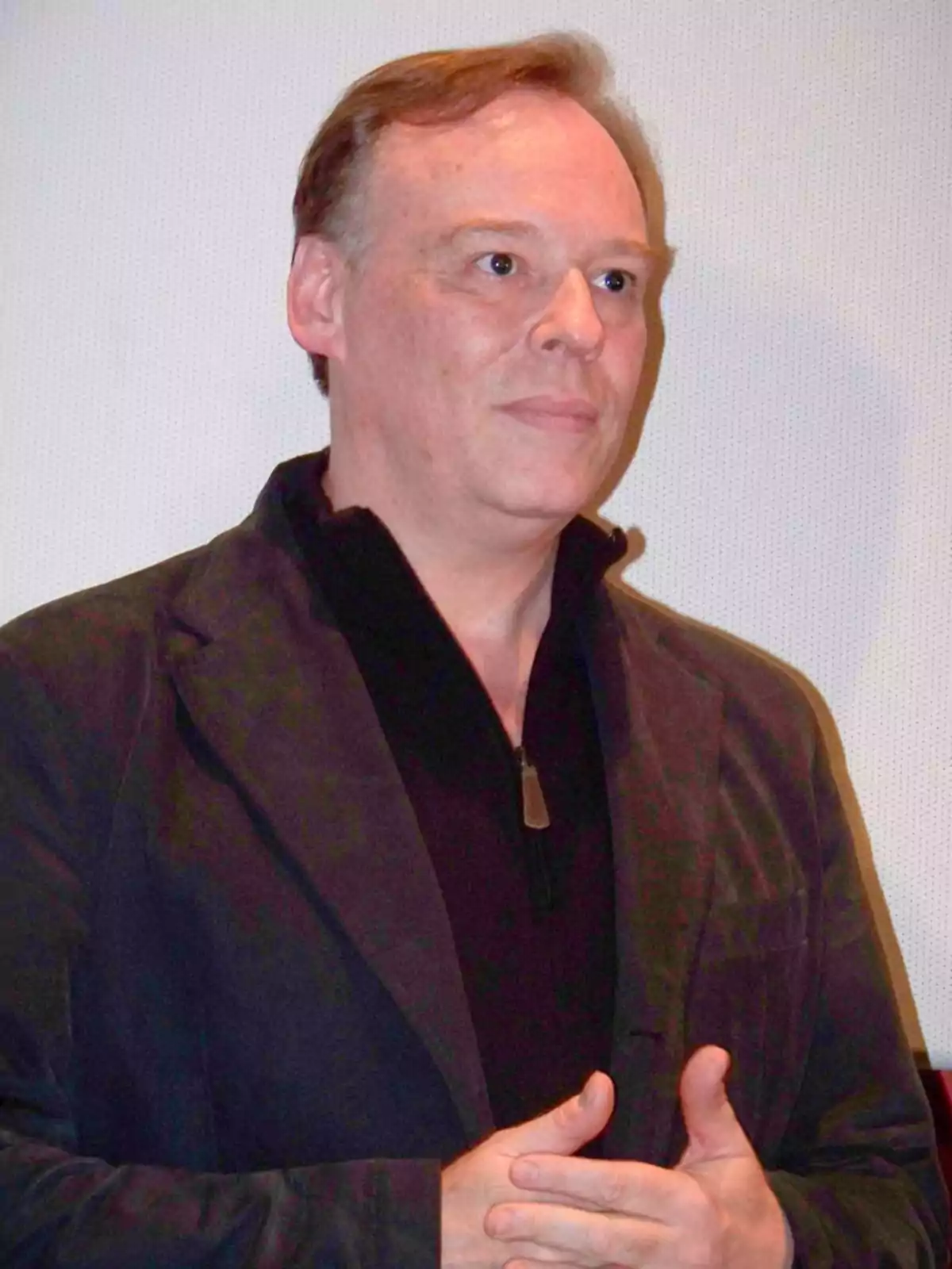Middle-aged man wearing a dark jacket and black jersey posing in front of a light-colored wall Middle-aged man wearing a dark jacket and black jersey posing in front of a light-colored wall
