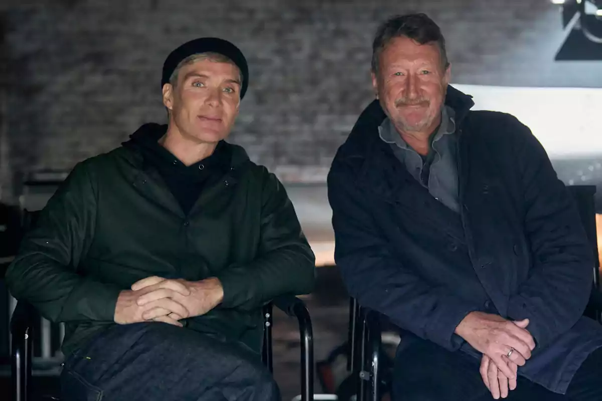 Two men sitting next to each other looking at the camera in an indoor setting with a blurred background Two men sitting next to each other looking at the camera in an indoor setting with a blurred background