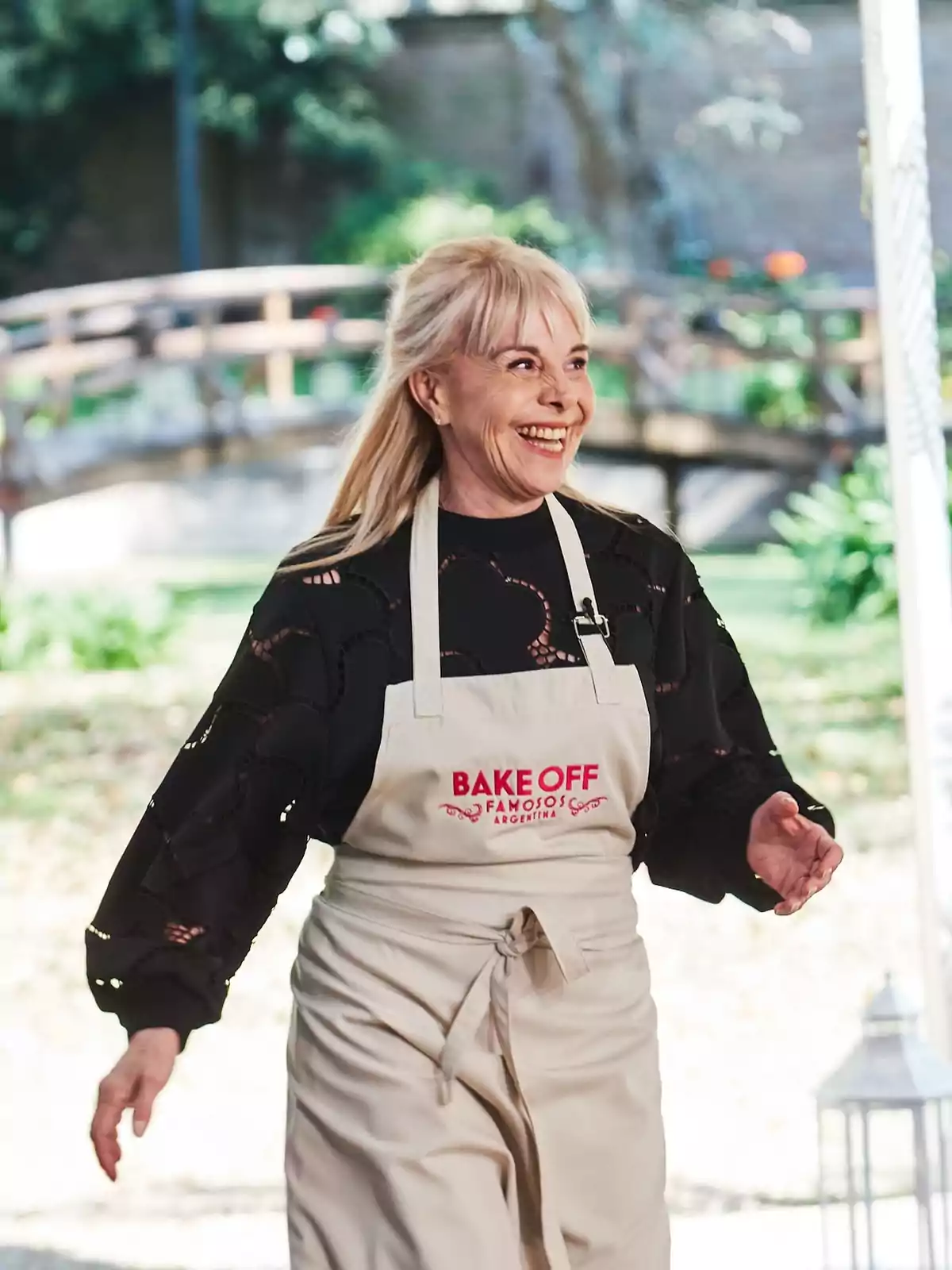Smiling blonde woman wearing a Bake Off Famosos Argentina apron in an outdoor setting with a wooden bridge in the background Smiling blonde woman wearing a Bake Off Famosos Argentina apron in an outdoor setting with a wooden bridge in the background