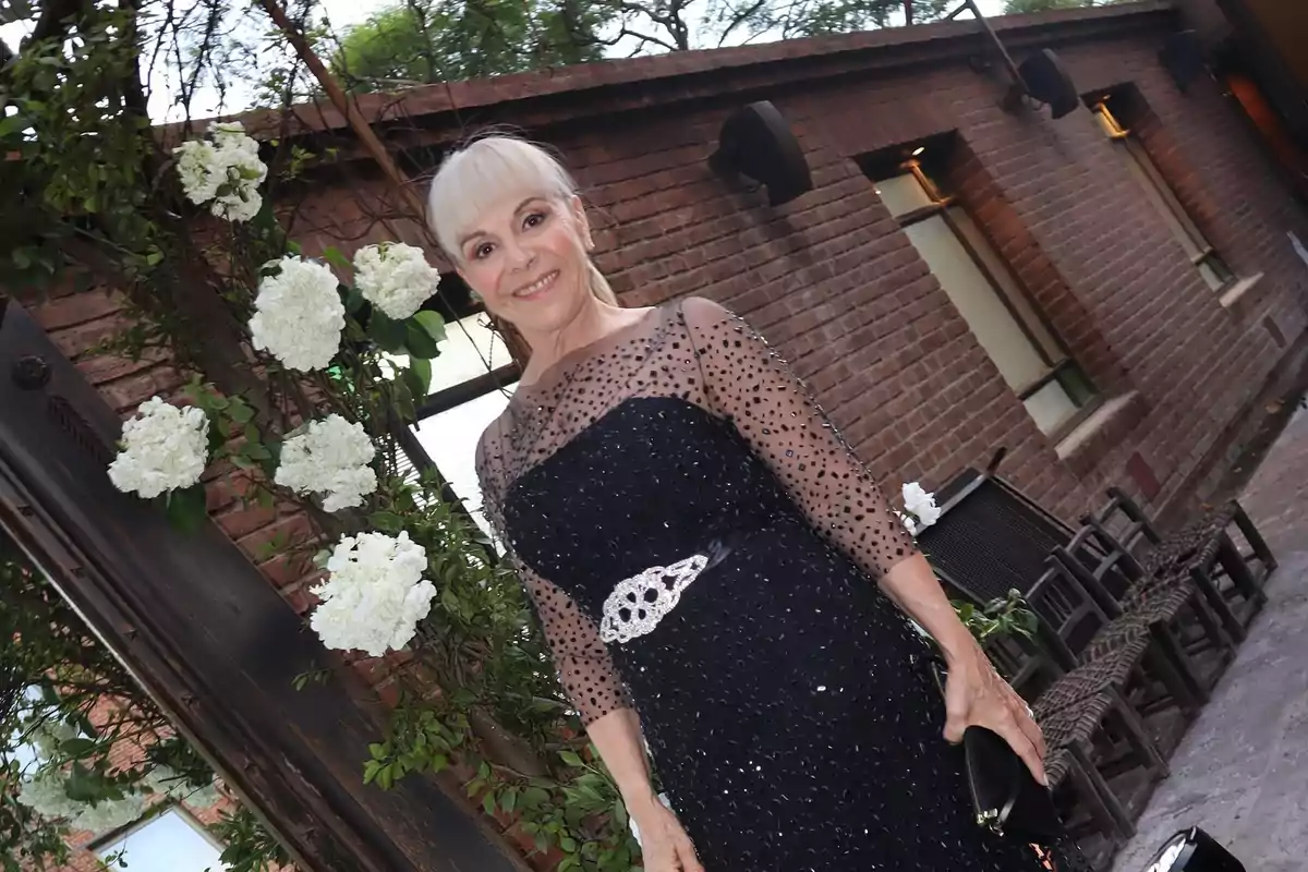 Smiling woman in an elegant black dress with sparkling details posing outdoors next to a brick wall and white flowers