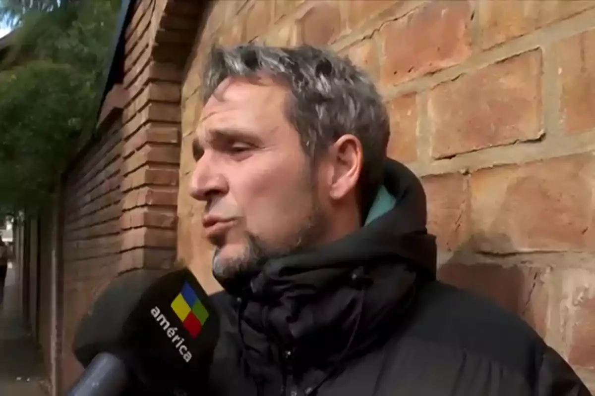Hombre de cabello canoso y barba corta siendo entrevistado por un canal de televisión frente a una pared de ladrillos