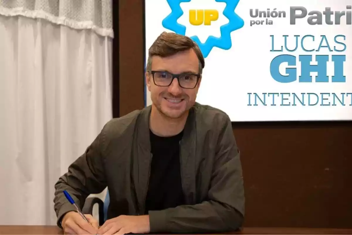 Hombre con gafas y chaqueta verde sentado en una mesa firmando un documento, al fondo un cartel de campaña política. Hombre con gafas y chaqueta verde sentado en una mesa firmando un documento, al fondo un cartel de campaña política.