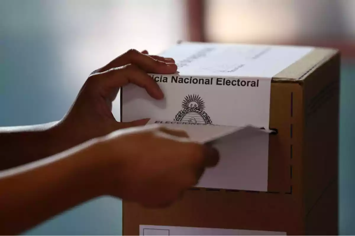 Mano introduciendo una boleta en una urna electoral de cartón con el texto Justicia Nacional Electoral y el escudo de Argentina. Mano introduciendo una boleta en una urna electoral de cartón con el texto Justicia Nacional Electoral y el escudo de Argentina.