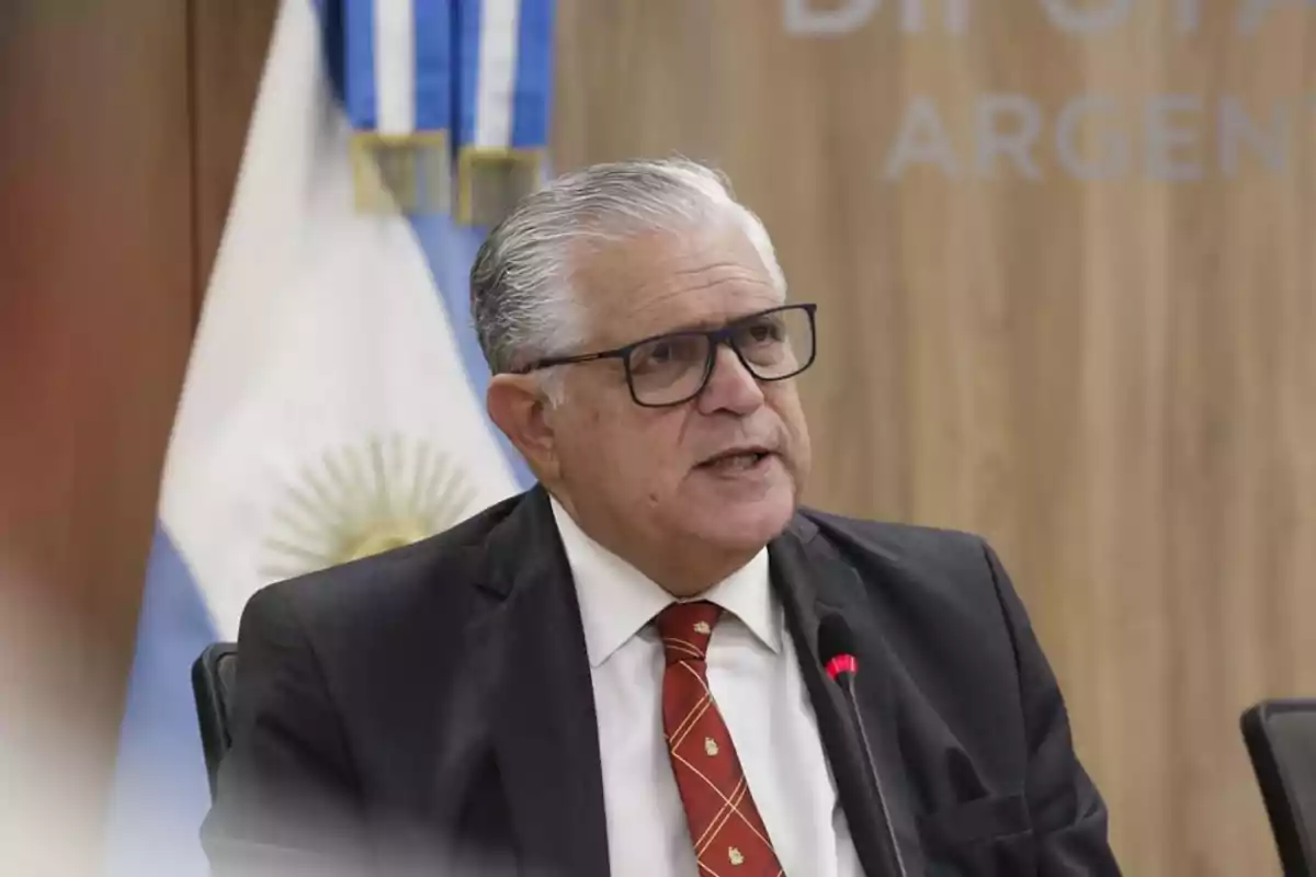 Hombre de cabello canoso y gafas hablando frente a un micrófono con una bandera de Argentina de fondo