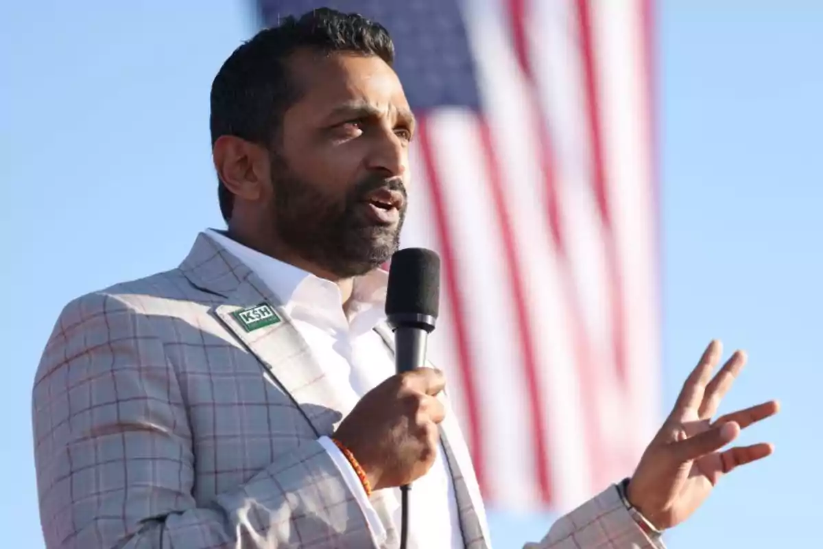 Bearded man speaking into a microphone, wearing a light plaid jacket, with a blurred United States flag in the background