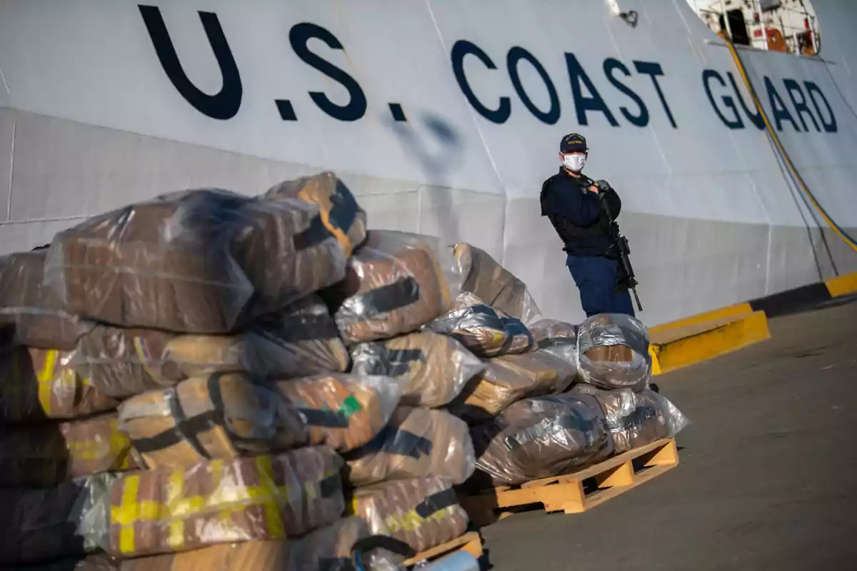 An armed Coast Guard officer wearing a mask stands next to numerous plastic-wrapped packages stacked on pallets in front of a United States Coast Guard vessel. An armed Coast Guard officer wearing a mask stands next to numerous plastic-wrapped packages stacked on pallets in front of a United States Coast Guard vessel.