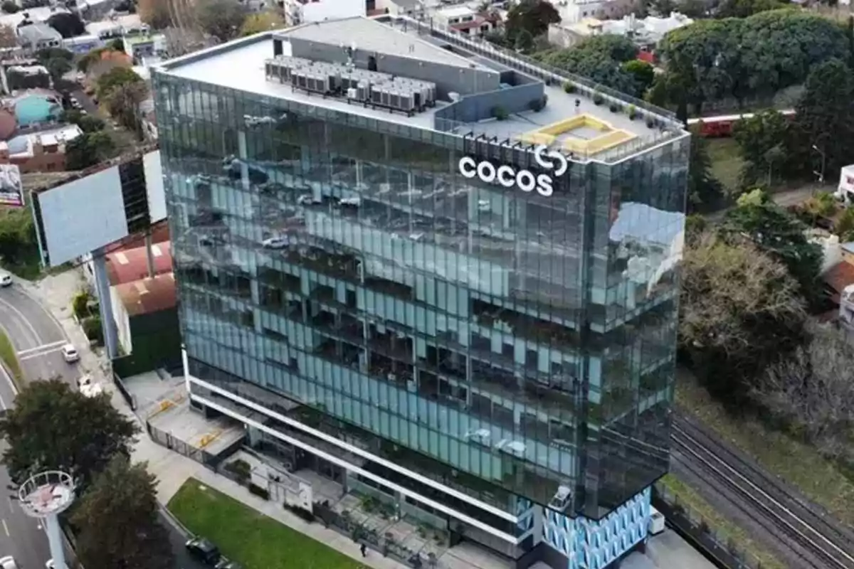Edificio moderno de oficinas con fachada de vidrio y el logo de COCOS en la parte superior rodeado de árboles y calles
