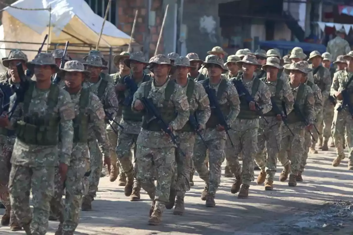 Soldados marchando en formación con uniforme de camuflaje y rifles en una calle