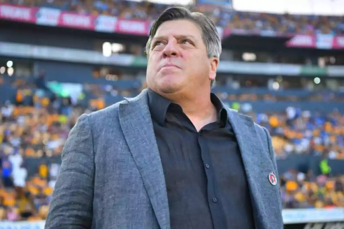 Hombre de cabello canoso con saco gris y camisa negra en un estadio lleno de espectadores