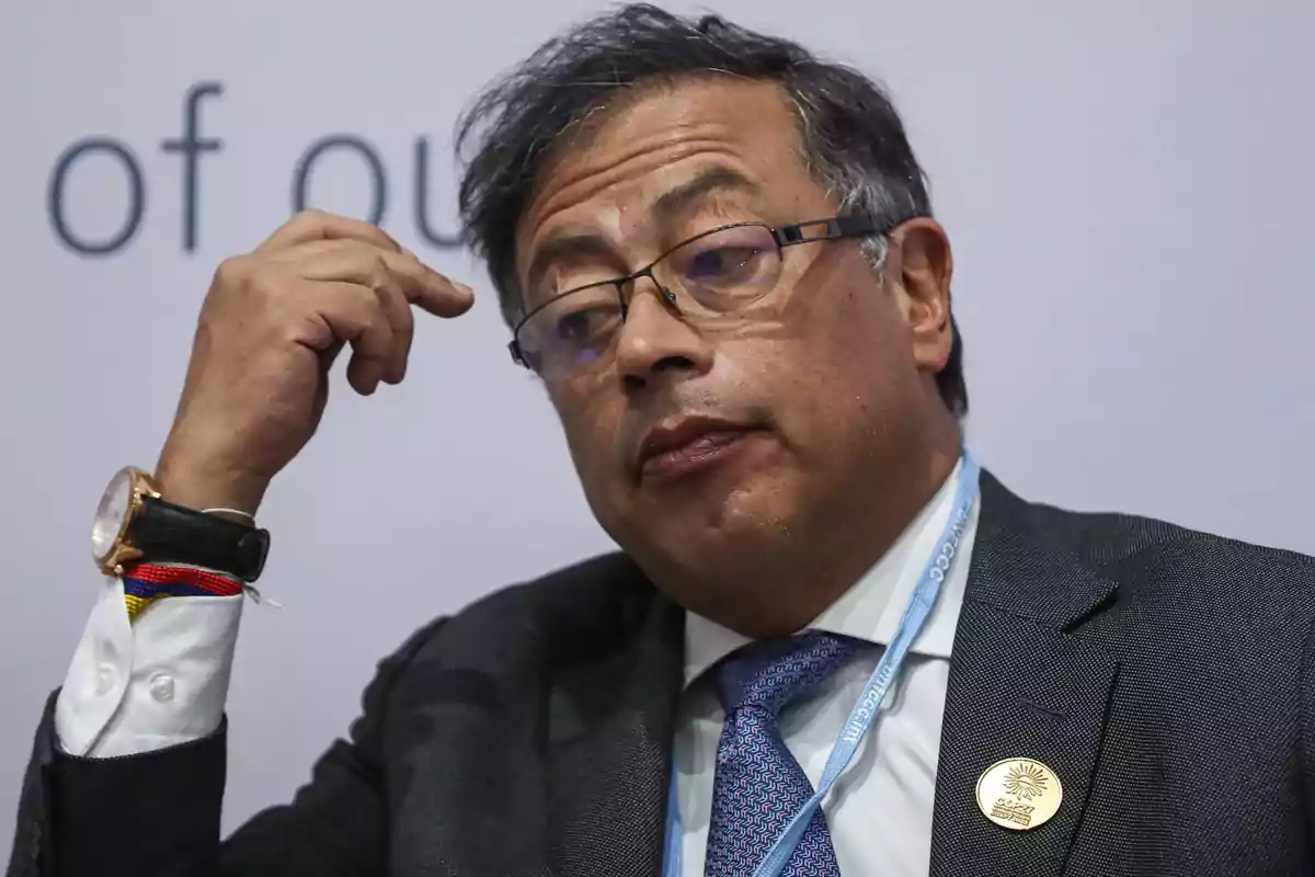 Hombre de cabello canoso y gafas con expresión seria, usando traje y corbata, tocándose la cabeza con una mano y portando un reloj con una pulsera de colores. Hombre de cabello canoso y gafas con expresión seria, usando traje y corbata, tocándose la cabeza con una mano y portando un reloj con una pulsera de colores.