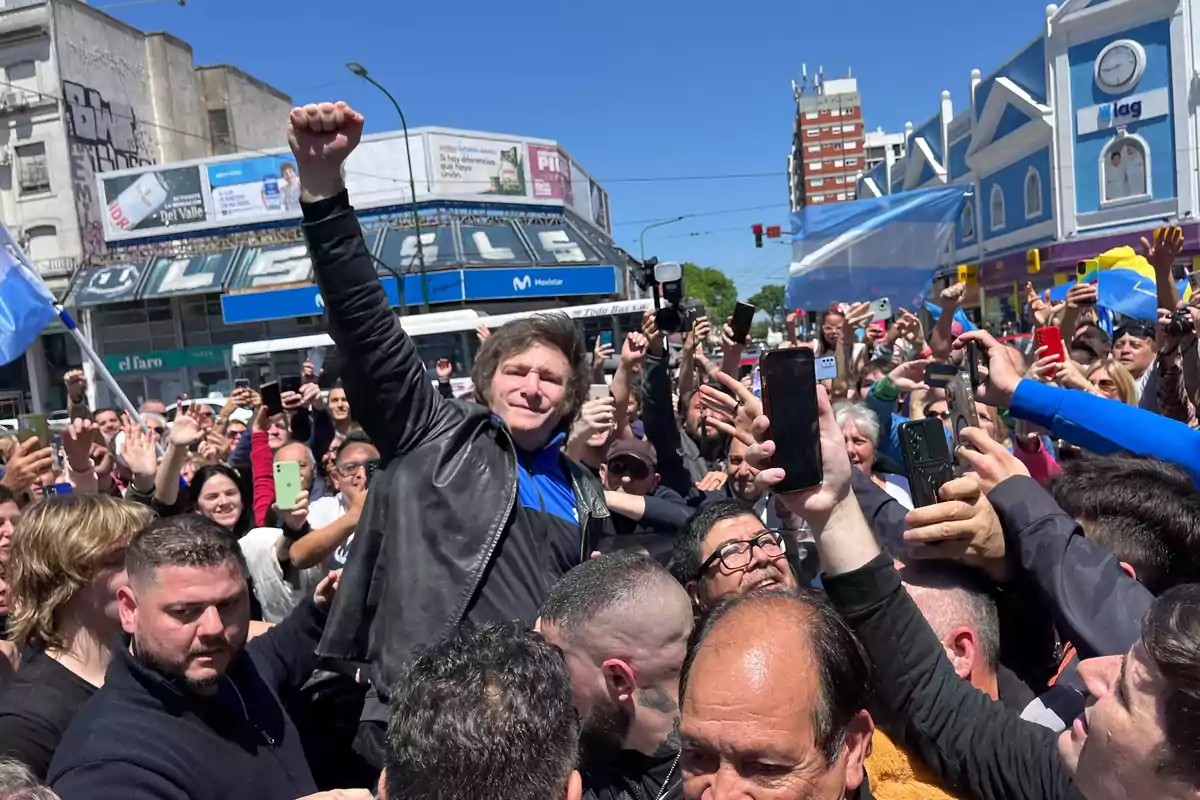 A man in a leather jacket raises his fist while a crowd surrounds him and takes photos on a sunny street with flags and buildings in the background. A man in a leather jacket raises his fist while a crowd surrounds him and takes photos on a sunny street with flags and buildings in the background.