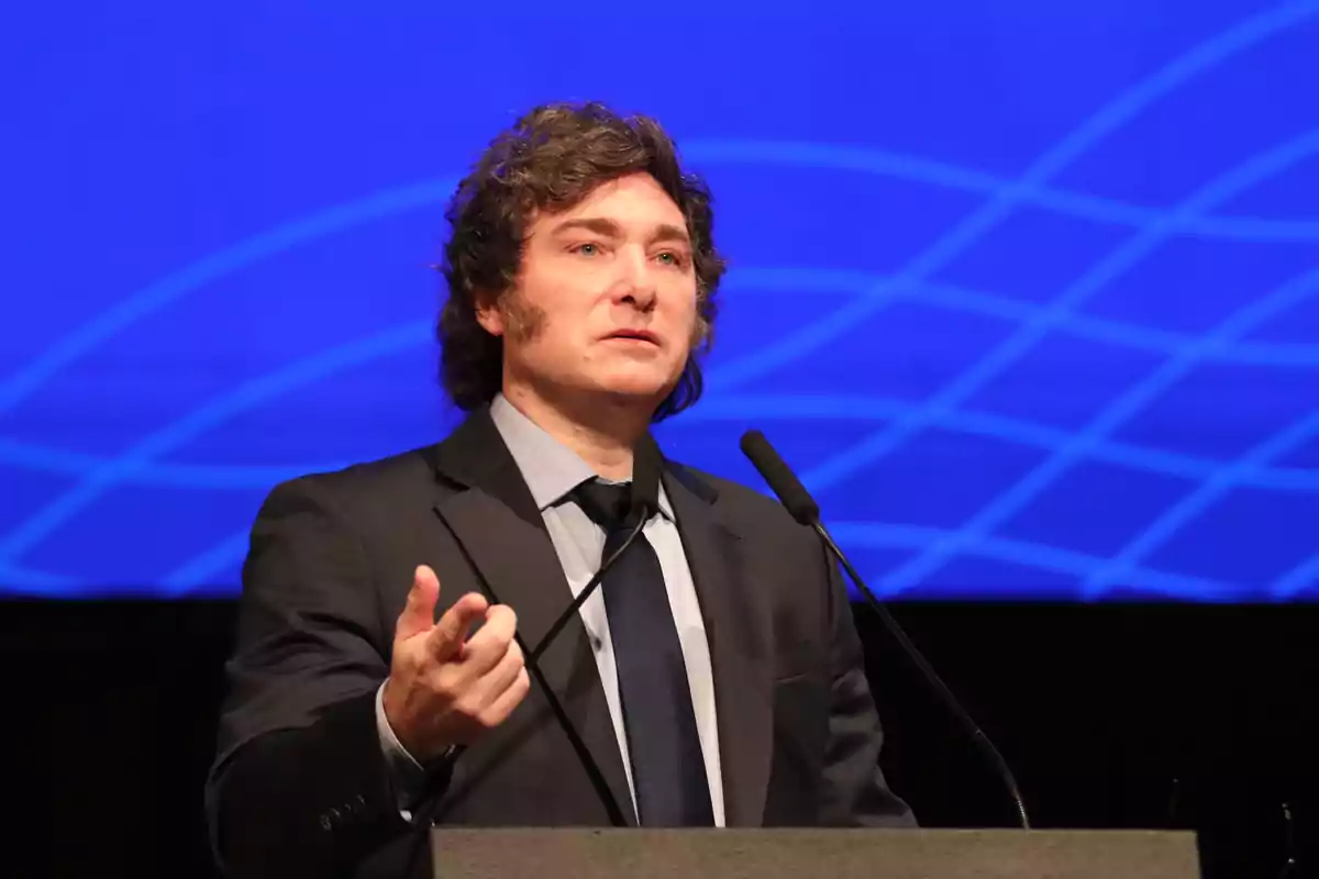 Curly-haired man in a dark suit speaking at a podium with a geometric blue background