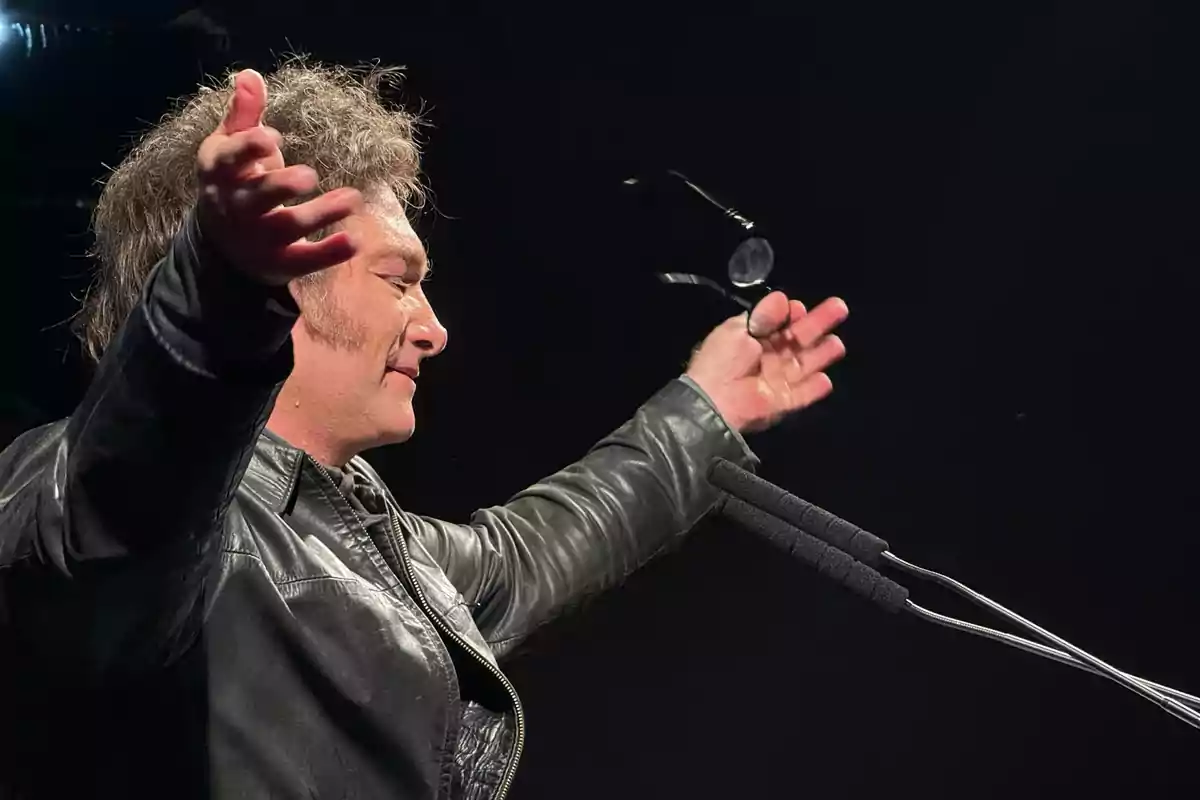 Man in a black leather jacket in profile, arms outstretched and holding glasses in front of a microphone against a dark background