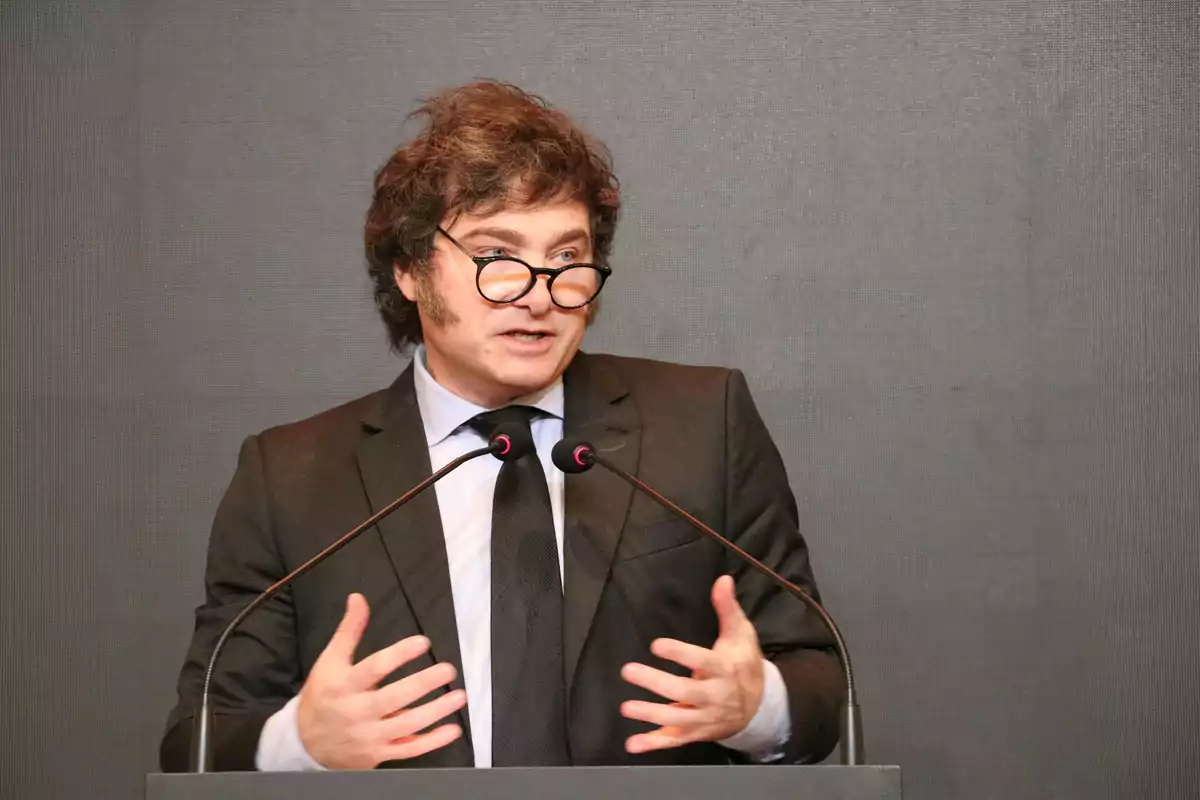 Man with glasses and a dark suit speaking at a podium with two microphones against a gray background