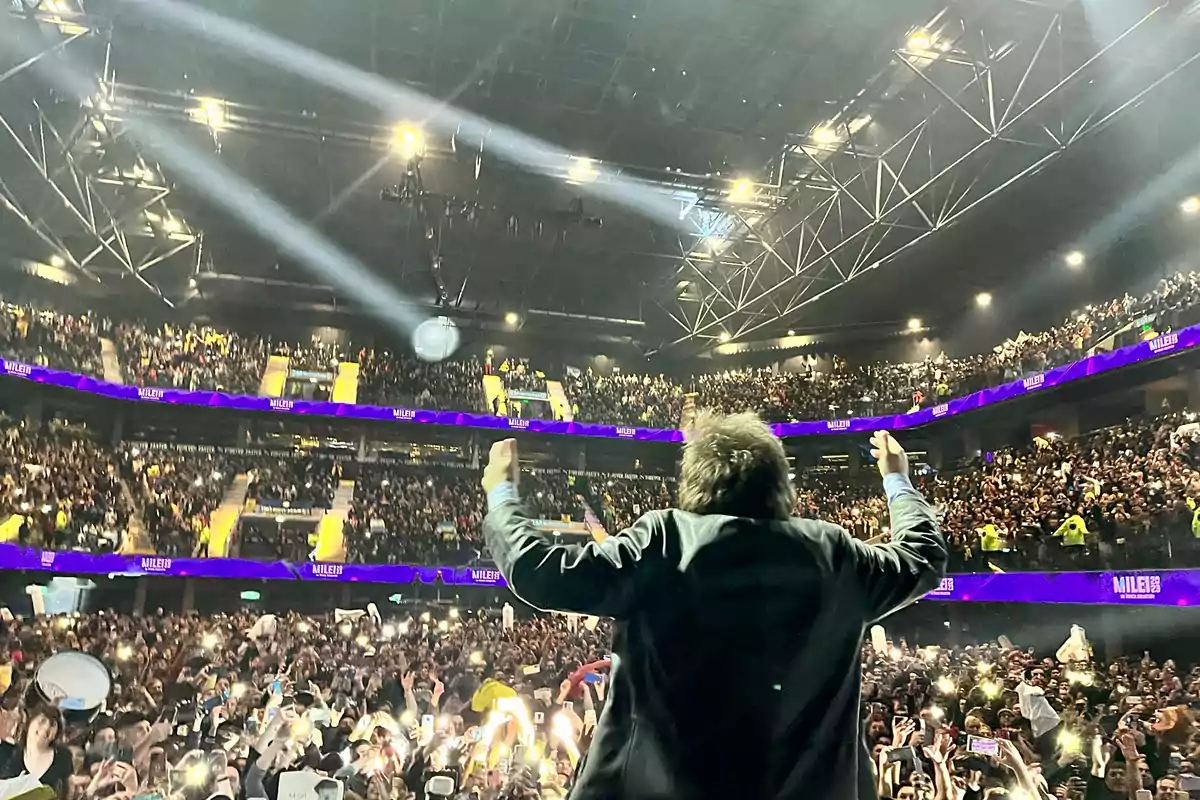 Persona de espaldas en un escenario frente a una multitud en un estadio iluminado con luces y pantallas moradas Persona de espaldas en un escenario frente a una multitud en un estadio iluminado con luces y pantallas moradas