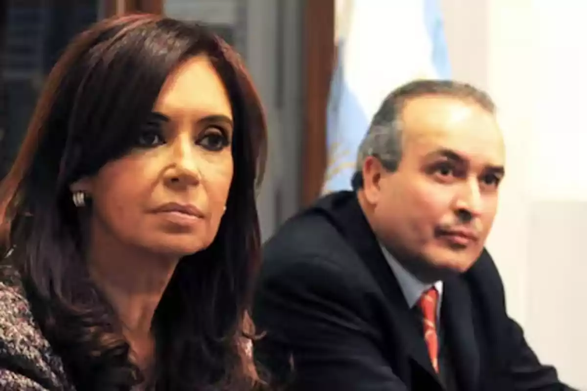 Dos personas sentadas en una mesa con expresión seria y una bandera de fondo