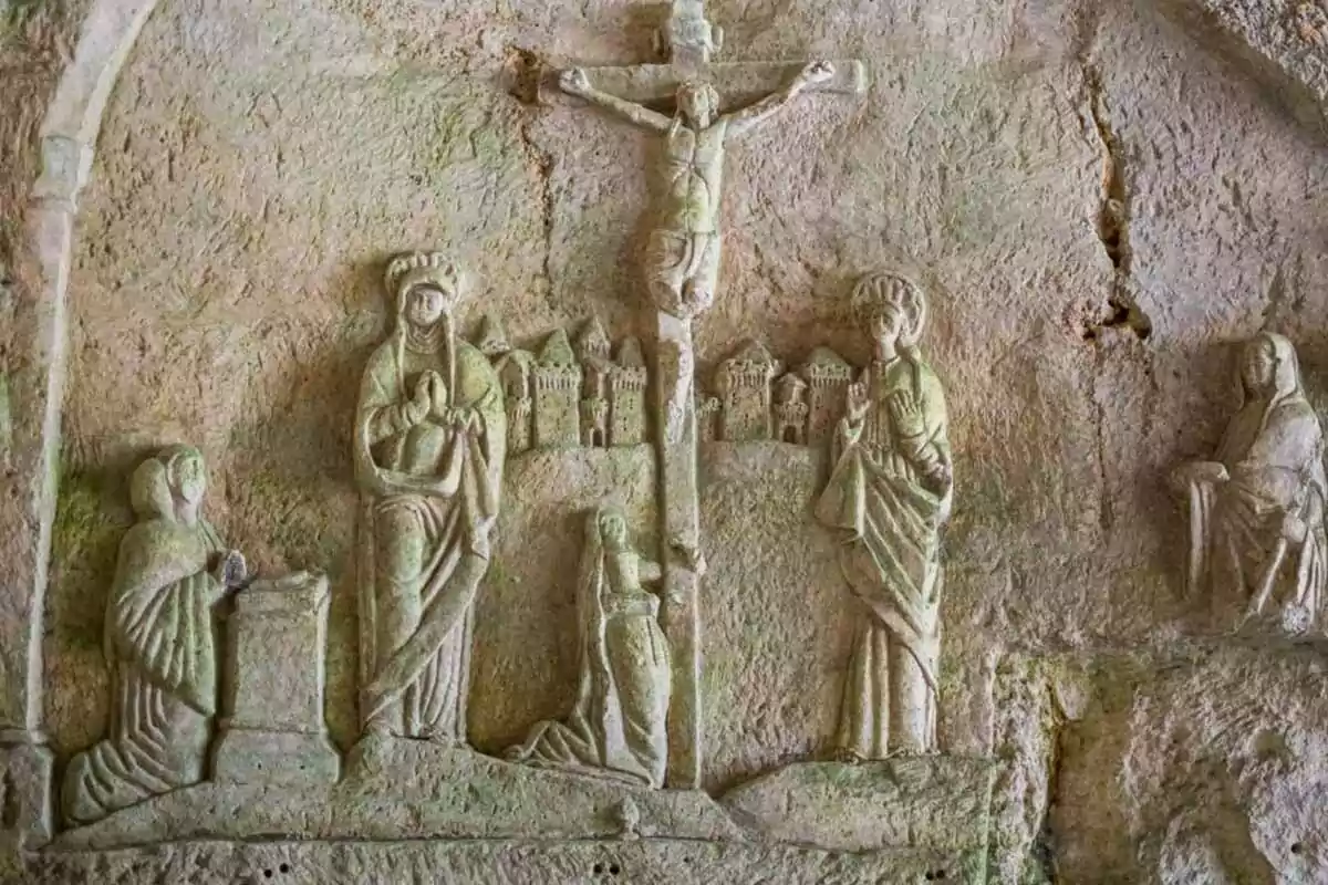 Relieve de piedra que representa una escena religiosa con figuras humanas y una figura central crucificada rodeada de otras personas en actitud de oración Relieve de piedra que representa una escena religiosa con figuras humanas y una figura central crucificada rodeada de otras personas en actitud de oración