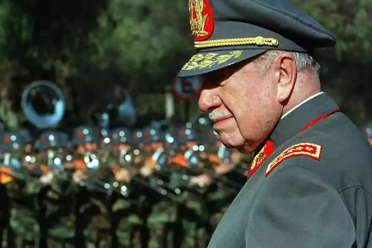 Un hombre de uniforme militar con gorra y charreteras doradas está de perfil en primer plano mientras un grupo de soldados desfila al fondo Un hombre de uniforme militar con gorra y charreteras doradas está de perfil en primer plano mientras un grupo de soldados desfila al fondo