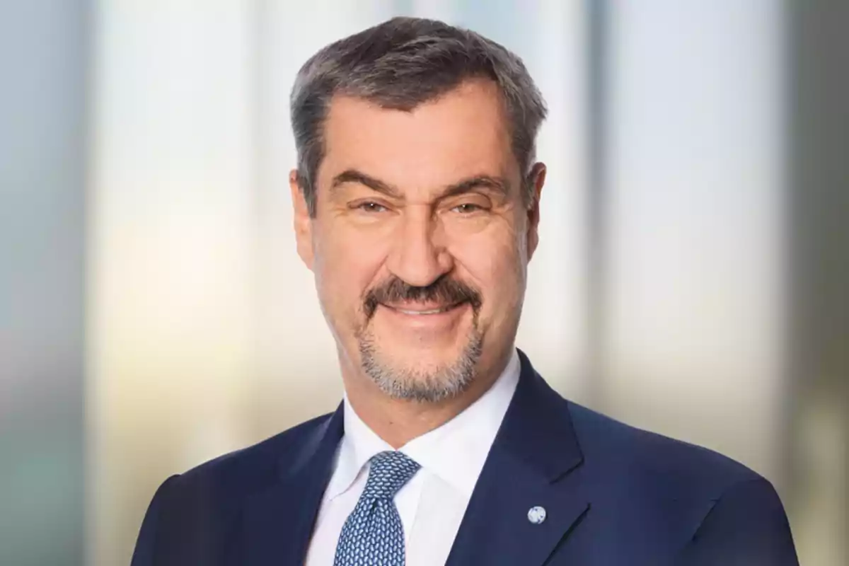 Middle-aged man with a beard and mustache, dressed in a blue suit and tie, smiling in front of a blurred background. Middle-aged man with a beard and mustache, dressed in a blue suit and tie, smiling in front of a blurred background.