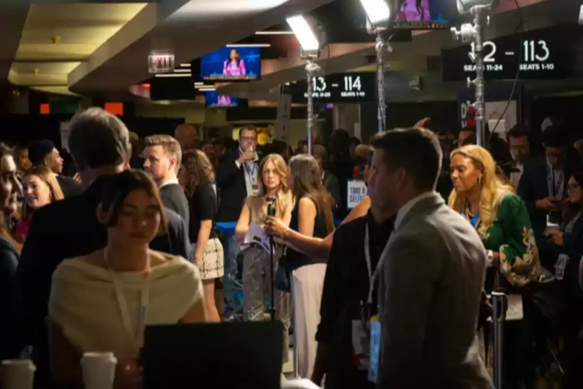 A group of people is gathered in a brightly lit indoor space; some are talking while others are observing, as screens and seat signs hang from the ceiling. A group of people is gathered in a brightly lit indoor space; some are talking while others are observing, as screens and seat signs hang from the ceiling.