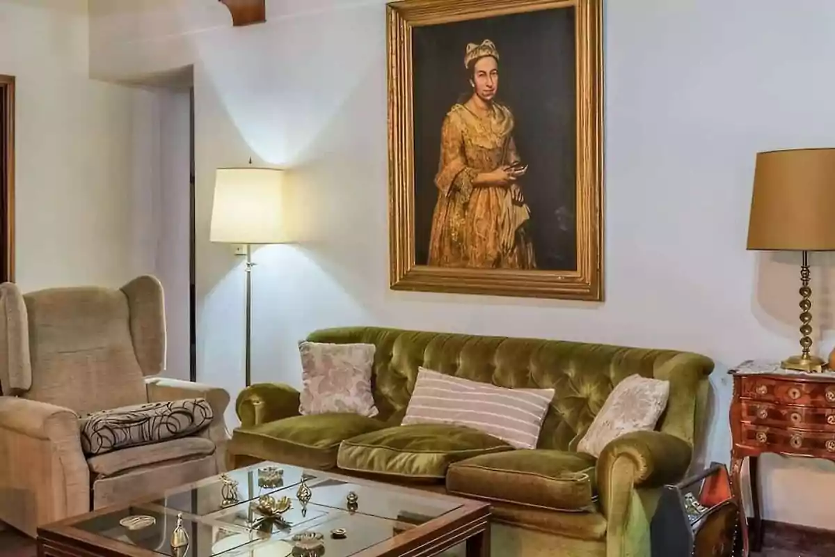 Living room with a green velvet sofa, beige armchair, glass coffee table, two lamps, and a large vintage portrait of a woman on the wall