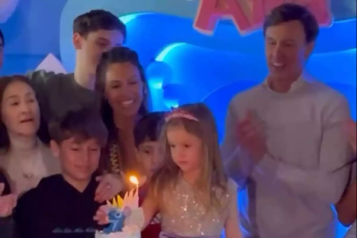 Un grupo de personas sonríe y celebra el cumpleaños de una niña pequeña que está frente a un pastel decorado con una vela encendida y una figura de Stitch