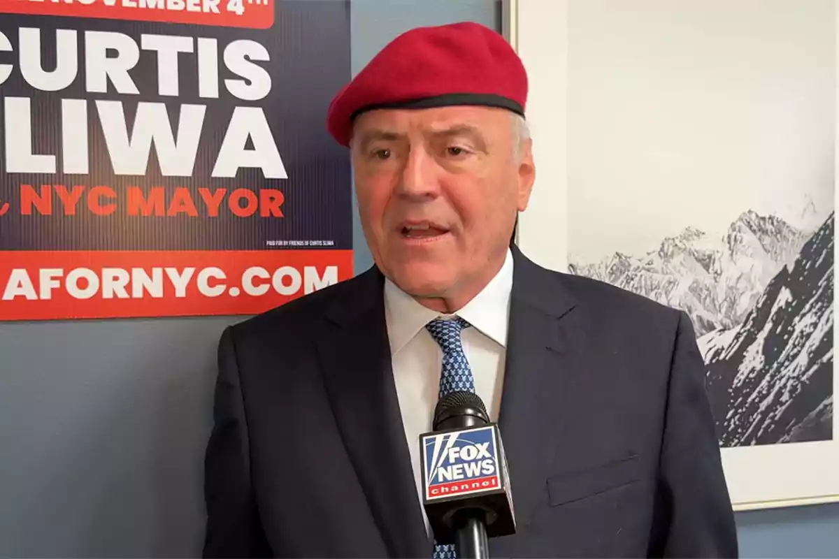Man wearing a red beret and dark suit speaking in front of a Fox News microphone with a political campaign sign and a photo of snow-capped mountains in the background Man wearing a red beret and dark suit speaking in front of a Fox News microphone with a political campaign sign and a photo of snow-capped mountains in the background